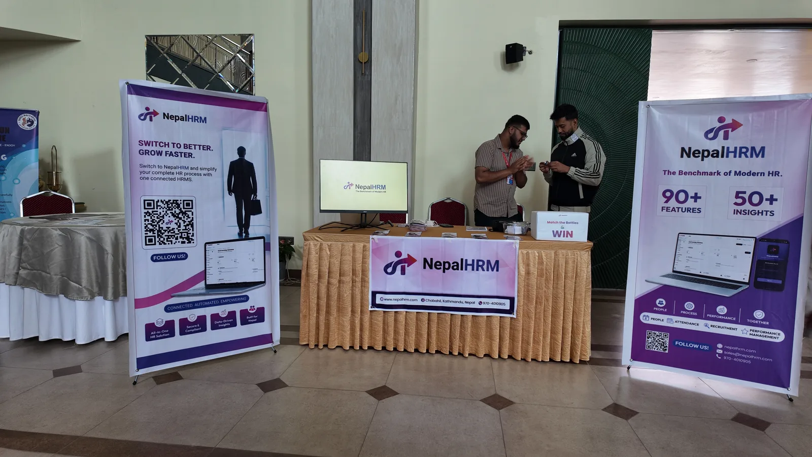 The finished NepalHRM stall at Idea Summit 2026: two standees listing the platform's modules, a branded table front, and a monitor running the product.