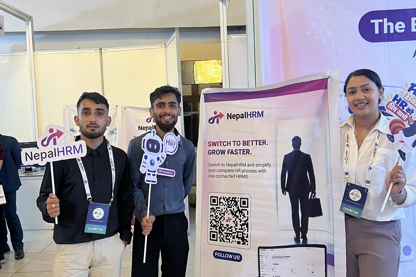 Two people at the NepalHRM stall holding a NepalHRM sign and a robot prop beside the roll-up banner, with a third on the right