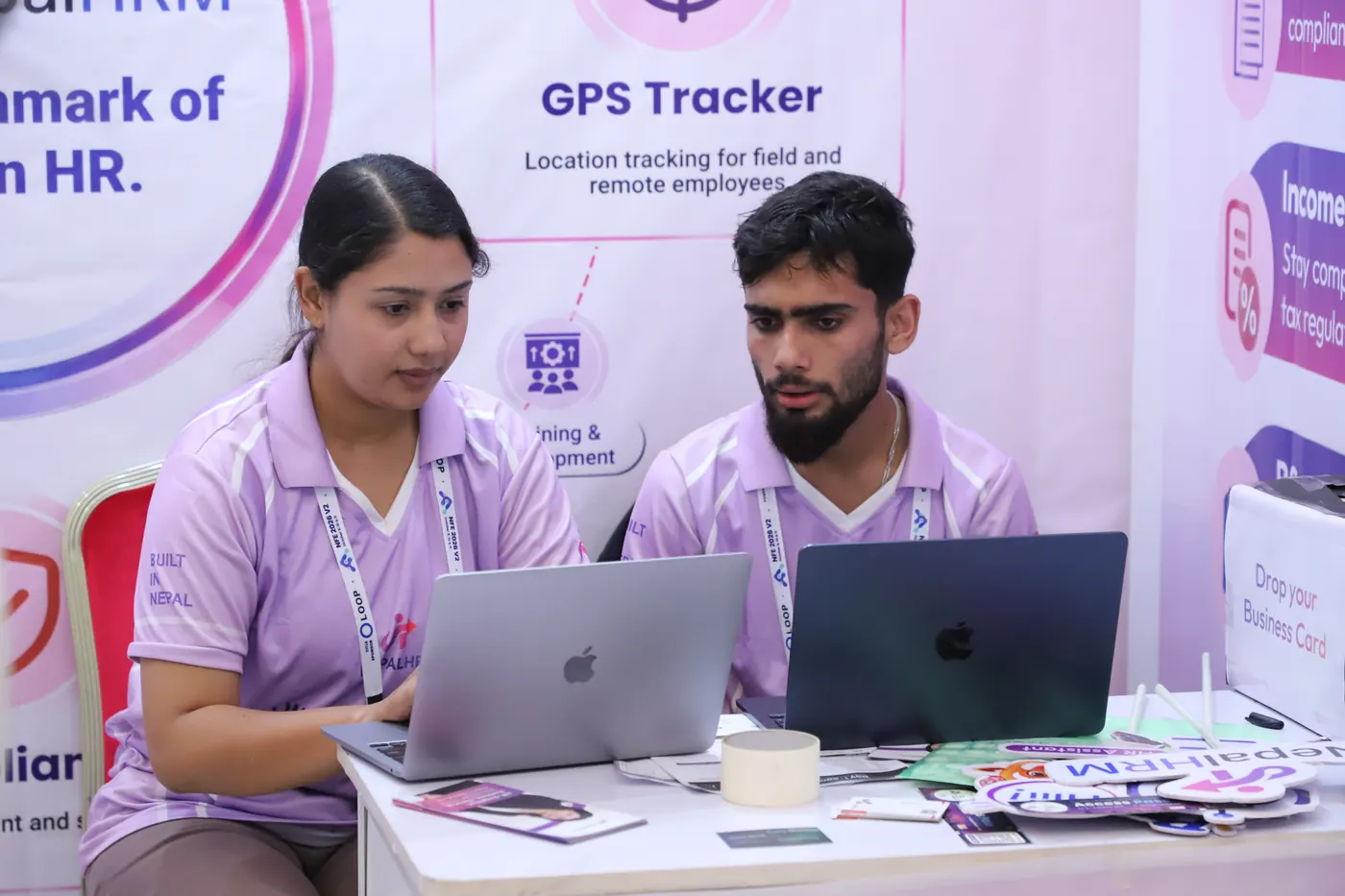 Two NepalHRM team members in lilac shirts working on laptops at the stall, in front of a panel about GPS tracking for field employees