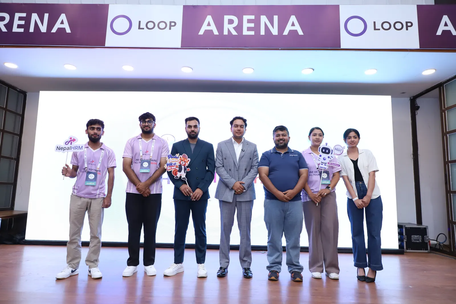 Seven people lined up on the Loop Arena stage after the NepalHRM pitch, holding a NepalHRM sign and a red panda mascot prop